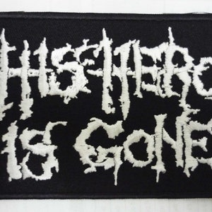 Op de afbeelding: Zwart rechthoekig embleem met witte geborduurde tekst "THIS HERO IS GONE" in een gotisch lettertype. De patch heeft een zwarte rand en is bedoeld voor kleding of accessoires.