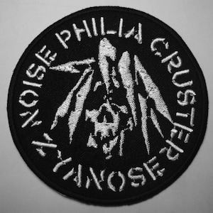 Peut inclure: Écusson brodé noir et blanc avec le texte "NOISE PHILIA CRUSTER" sur le bord. Un motif abstrait stylisé est au centre. L'écusson est rond.