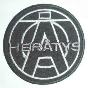Könnte beinhalten: Schwarz-weiß gestickter Patch mit einem stilisierten "A" in einem Kreis und dem Wort "HERATYS" darunter.