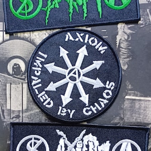 May include: Three black embroidered patches. The top patch is green and black with a dollar sign with a line through it, a creature with claws, and a circle with the letter A. The middle patch is black with a white circle containing a star with six points and a circle with the letter A in the center. The text around the circle reads "Axiom Impelled By Chaos". The bottom patch is white and black with a dollar sign with a line through it, a creature with claws, and a circle with the letter A.