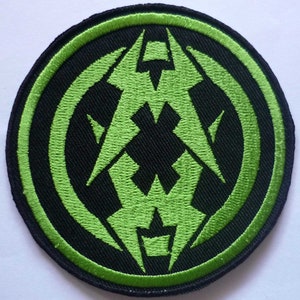 Könnte beinhalten: Runder schwarzer Aufnäher mit einem neon-grünen gestickten Design. Das Design zeigt eine zentrale geometrische Form mit eckigen Linien und einem kreisförmigen Rand. Der Aufnäher hat einen grünen Rand.