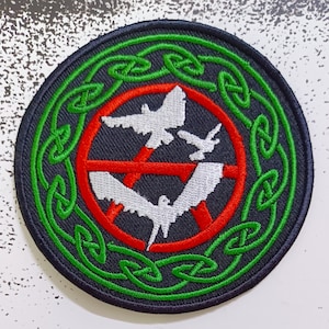 Può includere: Una toppa rotonda nera con un bordo di nodo celtico verde. All'interno del bordo c'è un cerchio rosso con un simbolo di anarchia bianco e due colombe bianche che volano attraverso il simbolo.