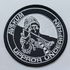 Puede incluir: Un parche bordado en blanco y negro con una persona con un gorro que sostiene un arma. El texto alrededor de la imagen dice "UPROR UNDERGRANZ AYSKUM".