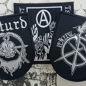 Peut inclure: Trois écussons brodés noirs avec des coutures blanches. L'écusson de gauche présente un crâne et le texte "disturd". L'écusson rectangulaire central montre un symbole anarchiste au-dessus de poings levés. Celui de droite affiche un oiseau et "PEACEFUL COLLAPSE".