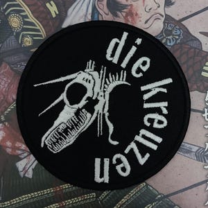 May include: A black and white embroidered patch with a skeletal creature and the text "die kruzen" in a circular design.