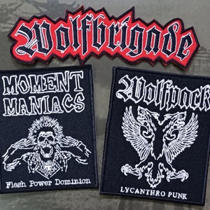 May include: Four black and white embroidered patches with red accents. The top patch reads "Wolfbrigade" in a gothic font. The left patch reads "Moment Maniacs" with a skull graphic and the text "Flesh Power Dominion". The right patch reads "Wolfpack" with a winged wolf graphic and the text "Lycanthro Punk".