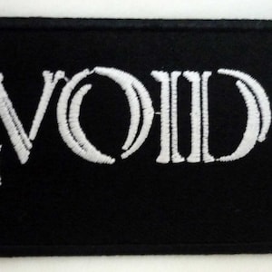 Pode incluir: Patch retangular preto com texto e símbolos bordados em branco. A palavra "VOID" está centrada, ladeada por uma cruz invertida à esquerda e uma cruz padrão à direita. Ideal para roupas ou acessórios.