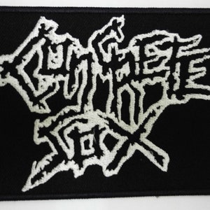 May include: Black rectangular patch with white embroidered text. The text appears to be a band logo or design, featuring stylized, jagged lettering. The patch has a black border and is likely made of fabric.