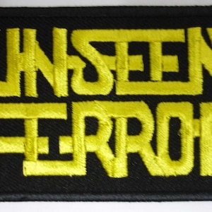 Op de afbeelding: Geborduurde patch met de woorden "UNSEEN TERROR" in gele draad op een zwarte achtergrond. De tekst is in een vet, gestileerd lettertype. De patch is rechthoekig.