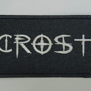 May include: A rectangular black embroidered patch featuring the white text ACROSTIX in a stylized font. The letters are stitched directly onto the black fabric background, which has a visible woven texture and a stitched border around the edge.