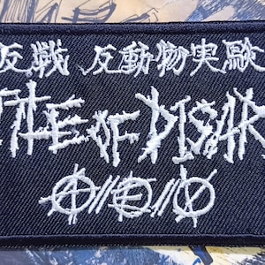 Op de afbeelding: Een zwarte geborduurde patch met witte letters die "反戦 反軍 侵略 実験" en "DISARM" lezen in een gestileerd lettertype. De patch heeft ook een cirkel met een diagonale lijn erdoorheen.