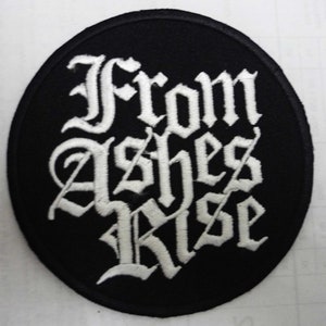 May include: A circular black patch featuring the embroidered phrase "From Ashes Rise" in white gothic lettering. The patch has a stitched edge and is designed for application on clothing or accessories.