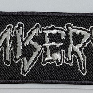 Peut inclure: Patch brodé noir avec le mot "MISERY" en blanc, entouré de deux symboles blancs.