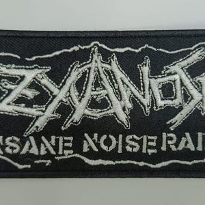 Może przedstawiać: Prostokątna czarna naszywka z wyhaftowanym białym napisem ZYANOSE wykonanym poszarpaną, stylizowaną czcionką nad słowami INSANE NOISE RAID zapisanymi blokowym, postarzanym krojem pisma, całość otoczona cienką białą obwódką wykonaną ściegiem.