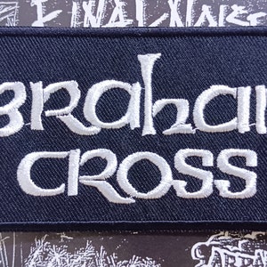 Peut inclure: Un écusson rectangulaire bleu marine brodé avec les mots "ABRAHAM CROSS" en blanc, dans une typographie stylisée. Ce patch est probablement destiné aux vêtements ou accessoires, et le design suggère un groupe ou une marque.
