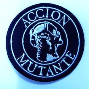 May include: A circular black patch featuring white embroidered text and a central design. The text reads "ACCION" above and "MUTANTE" below. The graphic is a detailed, stylized skull-like figure.