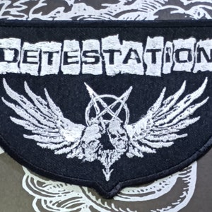 Op de afbeelding: Zwart-witte geborduurde patch met het woord "DETESTATION" in witte letters op een zwarte achtergrond. De patch heeft een witte gevleugelde schedel met een pentagram in het midden.