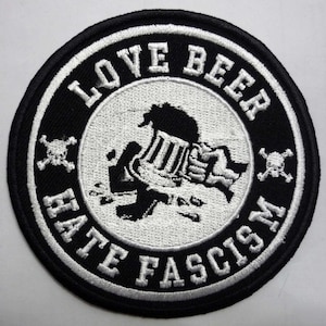 May include: Black and white embroidered patch with the text "Love Beer Hate Fascism" in a circle around a stylized image of a person holding a beer mug. Two skull and crossbones are on either side of the circle.