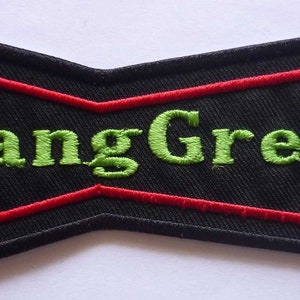 Puede incluir: Parche negro con borde rojo y el texto bordado "GangGreen" en verde. El parche tiene una forma única de pajarita, con un centro rectangular y lados angulados.