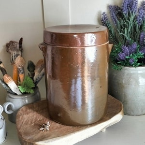 以下が含まれることがあります： 木製のトレイの上に置かれた、蓋付きの茶色の陶器の壺。壺は素朴でヴィンテージな外観で、控えめな釉薬で飾られています。トレイは天然木で作られており、ユニークな形をしています。