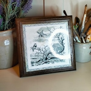 Op de afbeelding: Een ingelijste tegel met een zwart-wit ontwerp van een vogel die over een boom en een klein huis vliegt. De tegel is omgeven door een bruin houten frame.