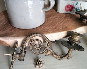 May include: A pair of ornate gold metal candle holders with a bee brooch on a light gray surface. The candle holders have a scroll design and are in the shape of a curved arm.