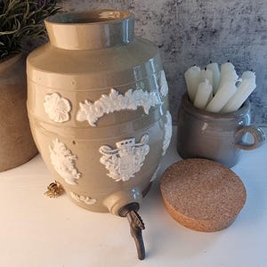 May include: A beige ceramic water dispenser with white floral and crest detailing. It has a spigot and is accompanied by a small mug with white candles and a cork stopper. The dispenser is approximately 38 cm tall.
