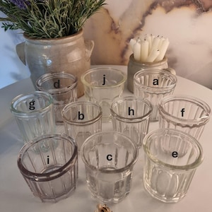 Könnte beinhalten: Eine Sammlung klarer Trinkgläser im Vintage-Stil mit geripptem Design. Die Gläser sind auf einer weißen Oberfläche angeordnet, zusammen mit einer kleinen dekorativen Bienenbrosche, einer Vase mit Lavendel und einem kleinen Topf mit weißen Kerzen.