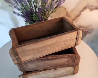 May include: Three stacked, weathered wooden boxes with metal corner reinforcements. The boxes are empty and have a rustic, aged appearance. A vase of purple lavender sits behind the boxes, adding a touch of color.