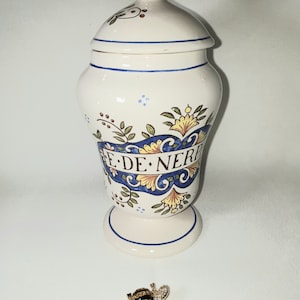 Pode incluir: Jarro de cerâmica branca com um design floral azul e dourado e o texto "E DE NERF". O jarro tem uma tampa e um broche em forma de abelha está em primeiro plano.