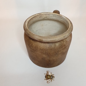 Peut inclure: Une tasse en céramique brune avec un intérieur blanc et une anse. Un petit bijou d'abeille en or se trouve au premier plan.