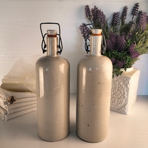Op de afbeelding: Twee vintage keramische flessen met metalen draadbevestigingen en kurken stoppen. De flessen zijn lichtgrijs van kleur met een gespikkelde afwerking. Ze staan op een wit oppervlak.