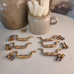 May include: A collection of vintage brass drawer pulls and hardware. The pulls have a curved design and are accompanied by matching screws. A small stoneware pot holds white candles. A larger stoneware pot is also present.