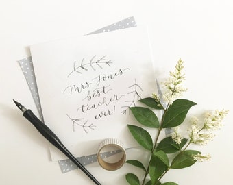 May include: A white card with the handwritten text "Mrs Jones best teacher ever!" is surrounded by a sprig of white flowers, a black pen, and a roll of decorative tape. The card is on a white surface with a gray polka dot paper.