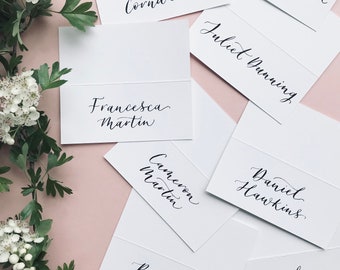 May include: White place cards with black calligraphy for a wedding or event. The names on the cards are Danny Cornwell, Francesca Martin, Sarah Foreman, Juliet Dunning, Cameron Martin, Daniel Hawkins, Brogan Lydonik, and Mark Zydonik.