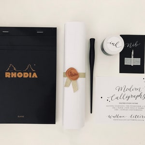 May include: A calligraphy kit with a black Rhodia notebook, a white envelope with a gold ribbon and wax seal, a black dip pen, a small jar of black ink, and a black card with the text "nib" and a grid pattern. The card also has a white card with the text "Modern Calligraphy" and "Wallace lettering" with a website address.