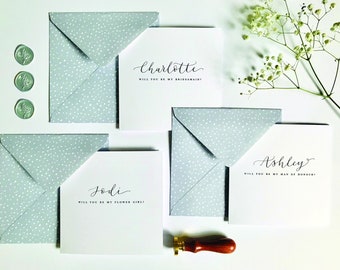 May include: Three white cards with gray envelopes, each with a different name and message. The cards say "Charlotte, Will you be my bridesmaid?", "Jodi, Will you be my flower girl?", and "Ashley, Will you be my man of honor?"