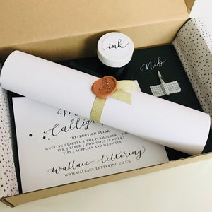 May include: A calligraphy kit in a brown box with white tissue paper. The kit includes a roll of paper, a black ink bottle, a nib, and a guide with instructions on how to get started with calligraphy.