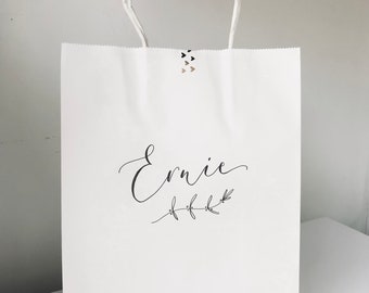 May include: White paper gift bag with a black handwritten name "Ernie" and a small black floral design.