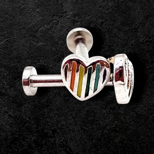 Puede incluir: Un labret de color plateado con un dije en forma de corazón. El corazón está relleno de rayas verticales en colores del arcoíris. El labret tiene una parte trasera plana y un poste roscado.