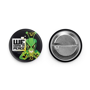 May include: A black and green button with a cartoon alien with a mischievous grin. The alien is holding its hands up in a gesture of peace. The text "WE COME IN PEACE" is written in white letters on a black background. The text "@shopboombaby by KID.ID" is written in smaller white letters below the main text.
