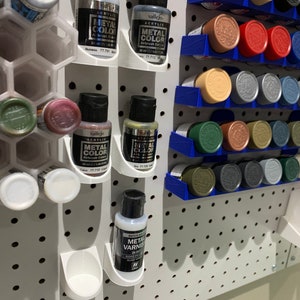 May include: A white pegboard with rows of small paint bottles in various colors. The bottles are organized in white and blue plastic holders. The bottles are labeled "Vallejo Acrylic Metal Color" and "Vallejo Acrylic Metal Varnish".