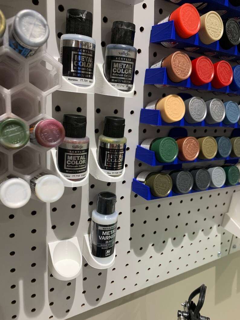 May include: A white pegboard with rows of small paint bottles in various colors. The bottles are organized in white and blue plastic holders. The bottles are labeled "Vallejo Acrylic Metal Color" and "Vallejo Acrylic Metal Varnish".