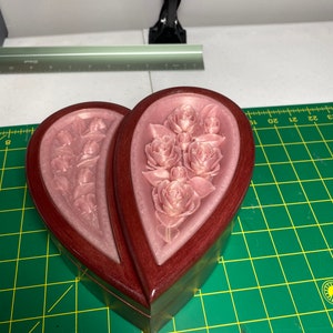 May include: A heart-shaped wooden box with a hinged lid. The box is made of dark wood with a light pink floral design on the inside. The box is closed and sitting on a green surface.