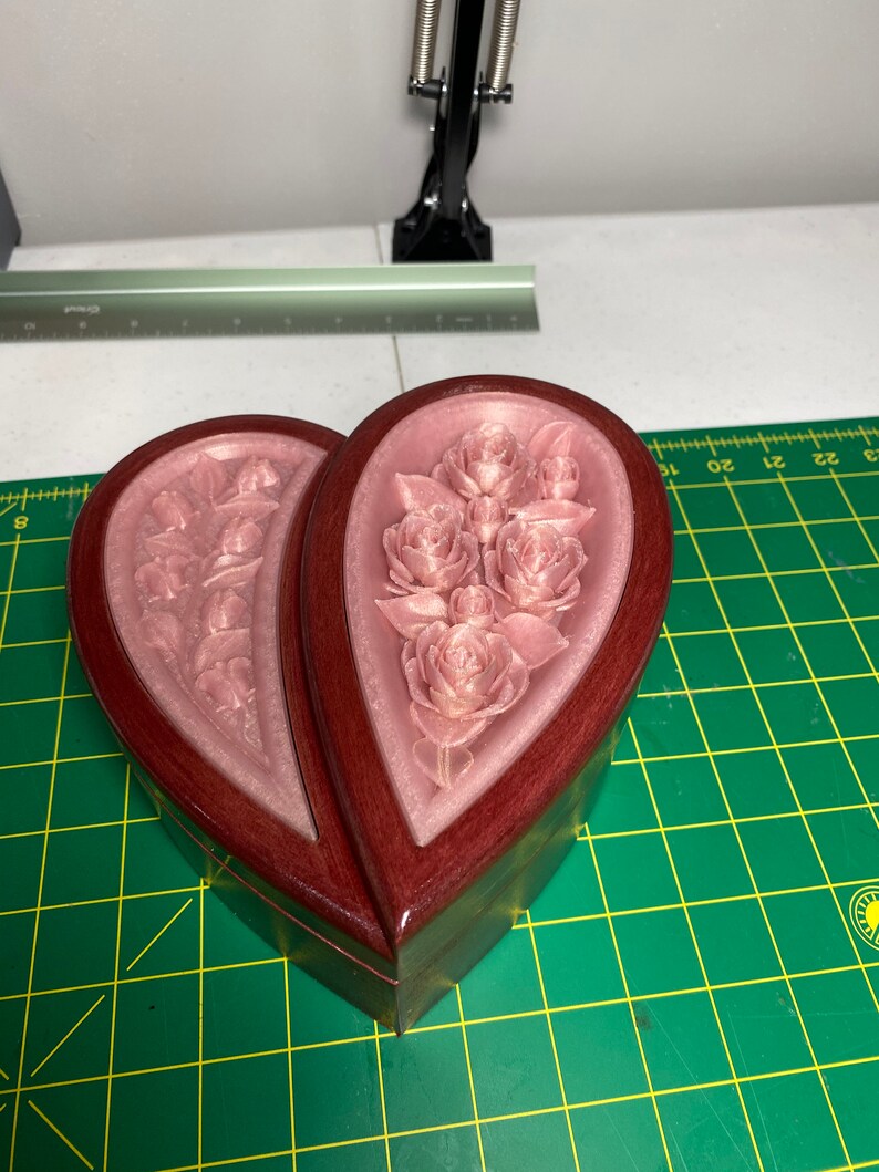 May include: A heart-shaped wooden box with a hinged lid. The box is made of dark wood with a light pink floral design on the inside. The box is closed and sitting on a green surface.
