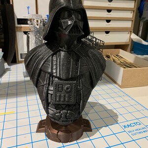 May include: A black 3D printed bust of Darth Vader from Star Wars. The bust is detailed and features a textured surface. The bust is sitting on a brown base.