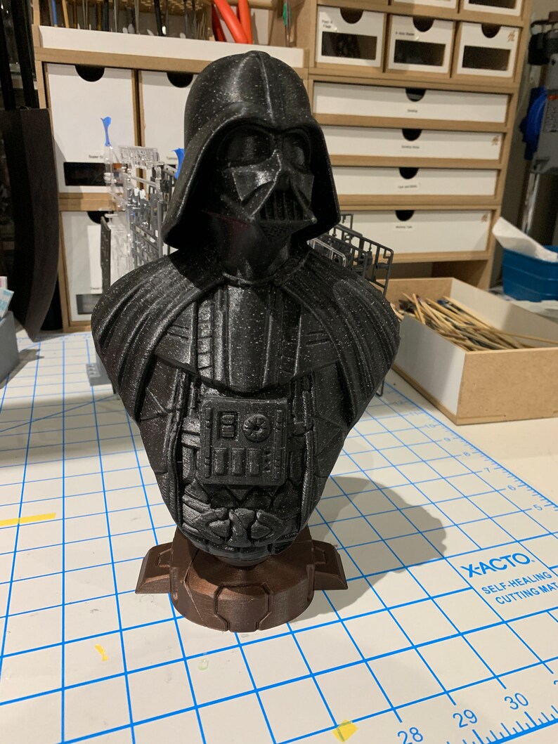 May include: A black 3D printed bust of Darth Vader from Star Wars. The bust is detailed and features a textured surface. The bust is sitting on a brown base.