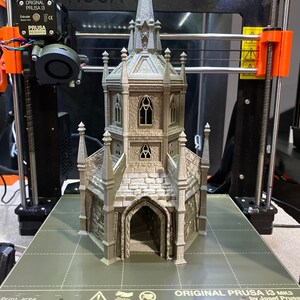 May include: A 3D printed model of a gothic chapel with a tall spire, intricate details, and a stone-like texture. The model is printed in a light gray color and is sitting on a black and gray textured surface. The text "ORIGINAL PRUSA i3 mk3 by Josef Prusa" is printed on the surface.