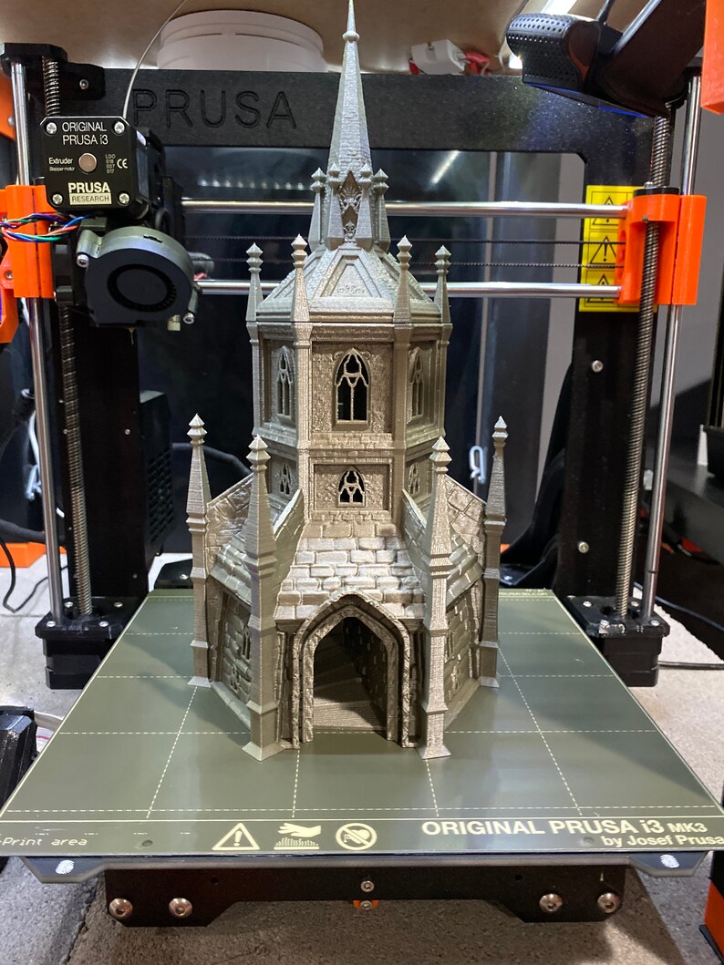 May include: A 3D printed model of a gothic chapel with a tall spire, intricate details, and a stone-like texture. The model is printed in a light gray color and is sitting on a black and gray textured surface. The text "ORIGINAL PRUSA i3 mk3 by Josef Prusa" is printed on the surface.