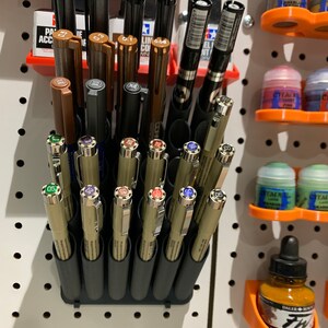 May include: A black plastic pen holder with 18 individual slots for pens and markers. The holder is mounted on a white pegboard wall. The holder is filled with a variety of pens and markers, including black, brown, and gold colored pens.